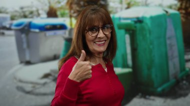 Kırmızı kazaklı, gülümseyen İspanyol kadın şehir sokaklarında renkli geri dönüşüm kutularının yanında, kentsel çevresel farkındalığın altını çiziyor..