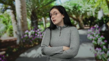 Kumral saçlı ve gözlüklü bir kadın açık hava parkında çiçeklerle ve doğal güzelliklerle çevrili dalgın bir ifadeyle duruyor..