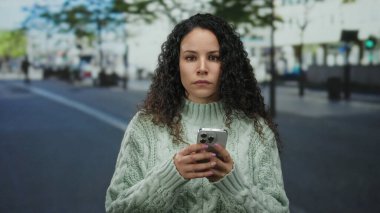 Açık şehir caddesinde akıllı telefon kullanan genç İspanyol kadın yeşil süveter giyiyor..