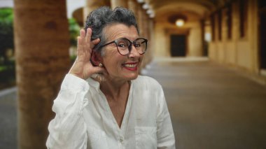 Gri saçlı yaşlı bir kadın, eli kulağında bir jest yapıyor, eski bir kasaba caddesinde gülümsüyor, şehir çevresi ile iç içe, açık bir atmosferi vurguluyor..