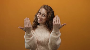 Turuncu arka planda gülümseyen bir kadın, rahat bir süveter içinde davetkar ve sıcak bir ortam sunan jestler yapar..