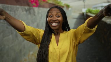 Kollarıyla gülümseyen kadın şehir sokağında sarı gömlekli, açık havada kıvırcık saçlı, gün içinde mutlu bir ifade sergiliyor..