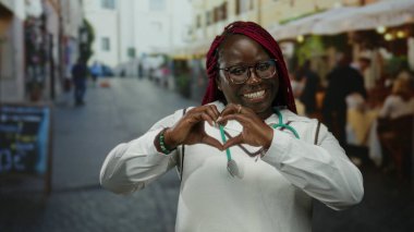 Steteskoplu kadın gülümsüyor ve kalp şekli oluşturuyor. Elleri şehir terasında, açık hava restoranında..