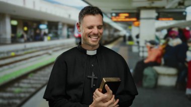 Tren istasyonunda elinde İncil 'le gezen rahip, gün içinde arka planda insanlarla gülümsüyor.