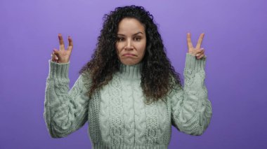 Yeşil kazaklı İspanyol bir kadın parmaklarıyla tırnak işareti yapıyor, mor bir arka plana karşı duruyor, belirsizlik ve iğnelemeyi ifade ediyor..