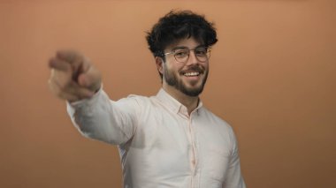 Beyaz gömlekli, sakallı, gözlüklü, turuncu arka planda poz veren, gülümseyen ve parmaklarını kafasında gezdiren genç bir İspanyol. Olumlu ve dostane bir hava yaratıyor..