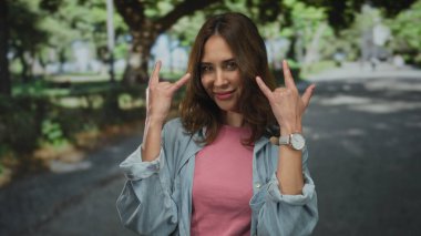 Genç İspanyol kadın güneşli bir parkta taş gibi jest yapıyor, sıradan bir kıyafet ve kot ceket giyiyor, kendine güvenen bir gülümsemeyle neşe ve yaratıcılık ifade ediyor..