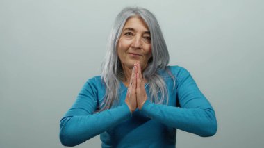Mavi tişörtlü yaşlı bir kadın izole edilmiş beyaz bir arka plana karşı el ele dua ederek huzur ve bilgeliği temsil eden huzur ve maneviyatı yayıyor..