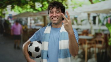 İspanyol bir adam sokakta kutlama yapıyor, elinde bir futbol topu tutuyor ve büyük bir gülümsemeyle parmaklarını sayıyor, spora ve canlı şehir atmosferine olan tutkusunu yakalıyor..