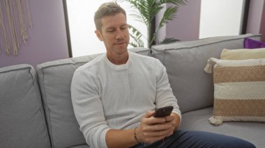 Genç adam rahat bir oturma odasında, şık dekorlar ve rahat yastıklarla çevrili akıllı bir telefon kullanarak koltukta dinleniyor.