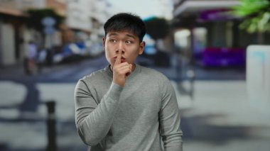 Genç adam hareketli bir şehir caddesinde sessizlik için el kol hareketi yapıyor, bir şehir ortamında entrika ve gizlilik anını yakalıyor..