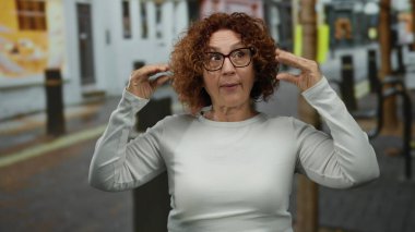 Orta yaşlı kıvırcık saçlı İspanyol kadın şehir sokaklarında gözlük ve beyaz gömlek takarak neşeli bir yüz ifadesi takınıyor..