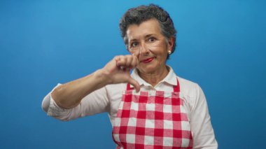 Önlük takan yaşlı bir kadın mavi arka plana karşı olumlu bir jest yapıyor. Ciddi bir ifadeyle memnuniyetsizliğini veya hoşnutsuzluğunu dile getiriyor..