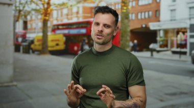 İspanyol adam kırmızı otobüsler ve binalarla çevrili bir şehir sokağında normal bir jest yapıyor, rahat giyiniyor ve kendine güvenini ve pozitifliğini ifade ediyor.