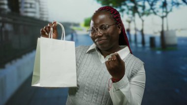Şehir merkezinde gülümseyen bir kadın, parlak bir günde, elinde alışveriş çantası ve parmaklarıyla davetkar bir şekilde el kol hareketi yapar..