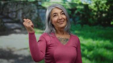 Pembe kazaklı, gri saçlı yaşlı bir kadın. Güneşli bir parkta gülümseyip el sallıyor..