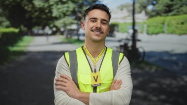 Yansıtıcı yelek giyen genç bir adam parkta gönüllü olarak çalışıyor, kendine güvenen bir ifadeyle kollarını kavuşturuyor, güneşli bir günde açık havada duruyor, bulanık yeşil bir arka planı var..