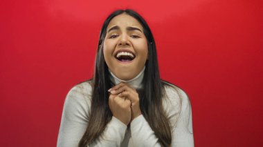 Ellerini sıkarak duygularını ifade eden, canlı kırmızı bir arka plana karşı duran, gerçek bir ifade sergileyen ve beyaz bir süveter giyen bir kadın..