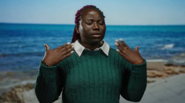 Elleriyle kendini yelleyen, deniz kenarında duran, yeşil kazak giyen, güneşli bir plaj gününde açık havada yaz esintisinin tadını çıkaran bir kadın..