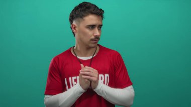 Kırmızı tişört giyen genç İspanyol cankurtaran, yeşil bir arka plan duvarına karşı düşünceli bir şekilde duruyor. İfadesinde ve duruşunda odak ve hazırlık gösteriyor..