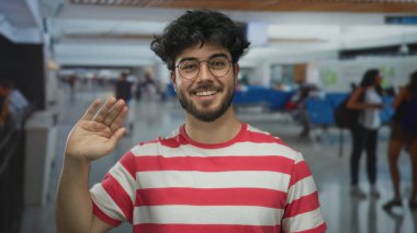 Sakallı ve gözlüklü genç bir adam, çizgili bir gömlek giyiyor, arka planda bulanık gezginlerle dolu kapalı bir havaalanında kameraya el sallıyor..