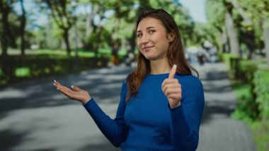 Mavi kazaklı genç kadın güneşli bir parkta, ağaçlarla çevrili ve bulanık insanları işaret ediyor..