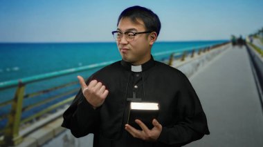 Dini giyinmiş genç adam deniz kenarında bir gezinti güvertesinde elinde arkasında okyanus olan bir kitap tutuyor..