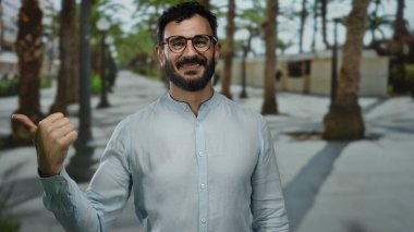 Dışarda duran sakallı İspanyol adam. Palmiye ağaçlarıyla gülümsüyor.