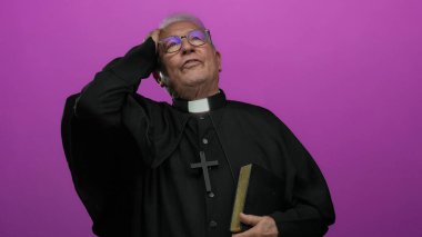 Siyah cüppeli kıdemli rahip, elinde bir kitap, pembe bir arka plan üzerinde duruyor, düşünceli bir ifadeyle, inancı ve düşünmeyi ön plana çıkarıyor..