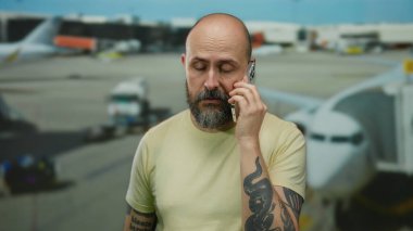 Havaalanında cep telefonu kullanan sakallı bir adam görünür ve açık hava atmosferi olan uçaklar, terminal ayarlarında seyahat ve iletişim sergiliyor.