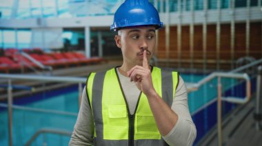 Güverte sandalyeleri ve denizle çevrili yolcu gemisinde sert şapka ve yansıtıcı yelek giyen genç adam susma hareketi yapıyor.