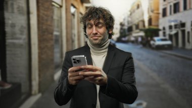 Şehir caddesinde akıllı telefon kullanan genç adam şık kıyafetler giyiyor odaklanmış ve içeriği kentsel mimari ve doğal ışıkla çevrili..