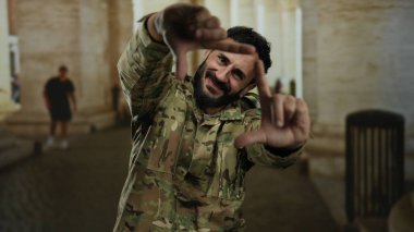 Askeri üniformalı İspanyol bir adam, eski Roma şehir ortamında gülümseyen elleriyle manzara çerçevesi çiziyor, hikaye anlatma ya da fotoğrafçılık öneriyor..