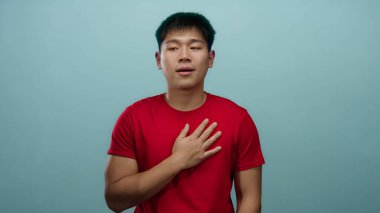 Kırmızı tişörtlü genç adam, elini göğsüne koyup mavi arka plan duvarına yaslanmış düşünceli ve içe dönük görünüyor..