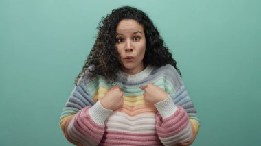 Latin bir kadın mavi arka plan duvarına karşı renkli bir süveter giymiş parmaklarıyla kendini işaret ederken şaşkın şaşkın bakıyor..