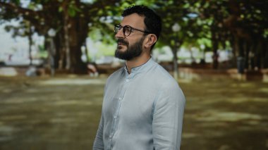 Sakallı İspanyol bir adam, güneşli bir parkta yeşil ağaçlar ve huzurlu bir ortam içinde, gözlüklü ve açık renk tişörtlü, sakin bir ifadeyi canlandırıyor..