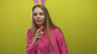 Tavşan kulaklı bir kadın, pembe gömlekli, sarı bir arka plana neşeli bir şekilde işaret ediyor. Genç, kaygısız bir havayla eğlenceli bir paskalya teması öneriyor..