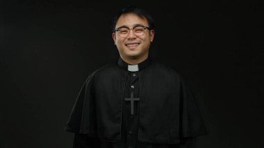 Gözlüklü ve siyah cüppeli Asyalı rahip karanlık bir arka plana gülümsüyor, el ele tutuşuyor ve dostça bir selamlaşma hareketi yapıyor gibi..