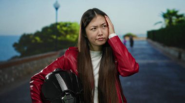 Kırmızı ceketli kadın miğfer tutuyor ve sahil kenarında düşünceli bir şekilde kafasına dokunuyor.