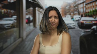 Şehir caddesinde duran genç kadın, bulanık trafik ve kentsel çevre ile şüpheciliğini dile getiriyor, açık hava atmosferi ve modern yaşam tarzını gözler önüne seriyor..