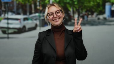 Açık havada gülümseyen bir kadın, günlük kıyafetleriyle şehir sokaklarında barış işareti yapıyor, ağaçların ve park halindeki arabaların olduğu canlı bir şehir sahnesini çekiyor..
