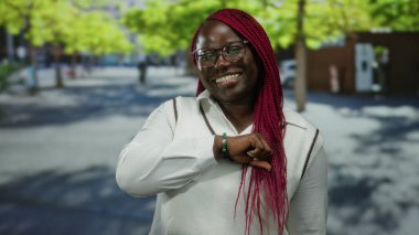 Şehrin sokak ortamında pembe saçlı ve gözlüklü bir kadın, güneşli bir günde kendine güven ve neşe gösteriyor, yeşil ağaçlar ve kentsel çevre ile çevrili..