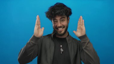 Sakallı İspanyol bir adam canlı mavi bir arka plan önünde coşkuyla jest yapıyor, etkileyici ve yaratıcı bir enerji sergiliyor..