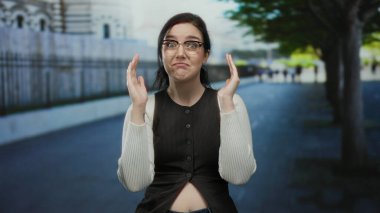 Açık havada esmer bir kadın, sokak sahnesinde ağaçlarla el hareketi yapıyor, gözlük takıyor ve gülümsüyor..