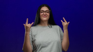 Gözlüklü kadın gülümsüyor ve mavi arka plana taş el hareketi yapıyor..