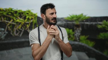 Sakallı İspanyol bir adam, yeşil ağaçlarla çevrili bir parkta dikilirken kaygılı görünüyor. Beyaz gömlek ve pantolon askısı takıyor..