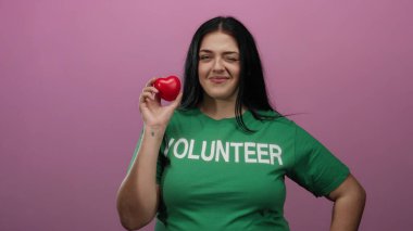Kırmızı kalp şeklinde bir nesne tutan kadın pembe arka plan aydınlatma ve nezaket karşısında gönüllü gömleği giyerek güvenle gülümsüyor..
