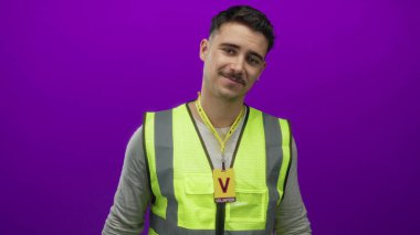 Yansıtıcı yelek ve gönüllü rozet giyen İspanyol asıllı genç adam izole edilmiş mor arka plan duvarına karşı duruyor, gençlik, gönüllülük ve kamu hizmeti hissi veriyor..