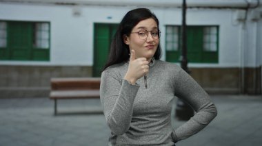 Esmer saçlı, gözlüklü bir kadın, arka planda yeşil kapılar olan şehir sokağında el kol hareketleri yapıyor, dinleme ve onay ifadeleri takınıyor..