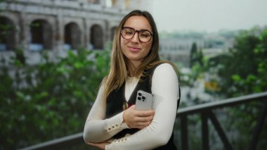 Roma Kolezyumu 'nun yakınlarında gülümseyen genç bir kadın aydınlık bir günde gözlük takıyor ve doğa arka planıyla çevrili şık bir kıyafet giyiyor..